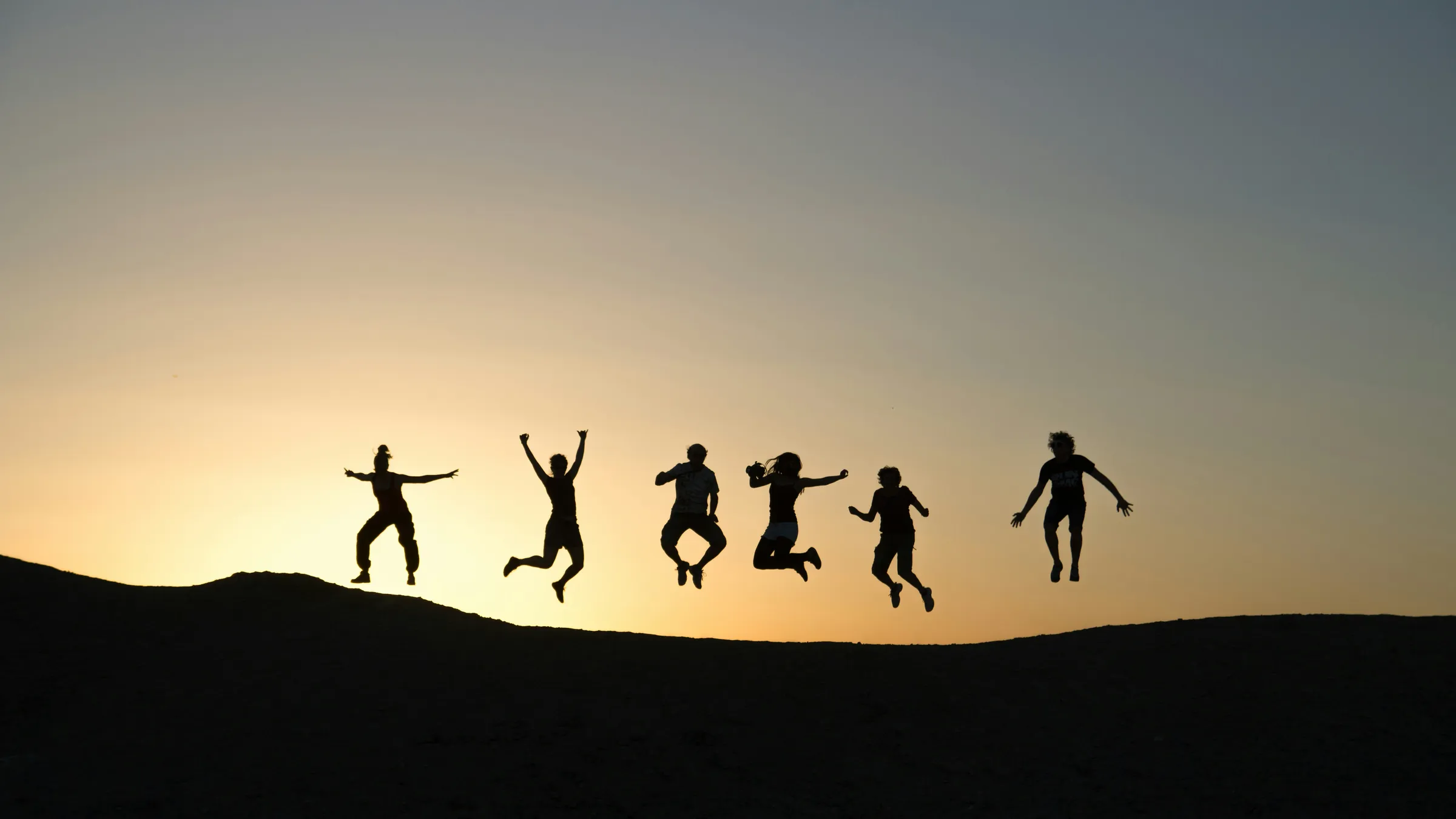 Six friends jump on a ridge at dusk, silhouetted against the last of the sunset.