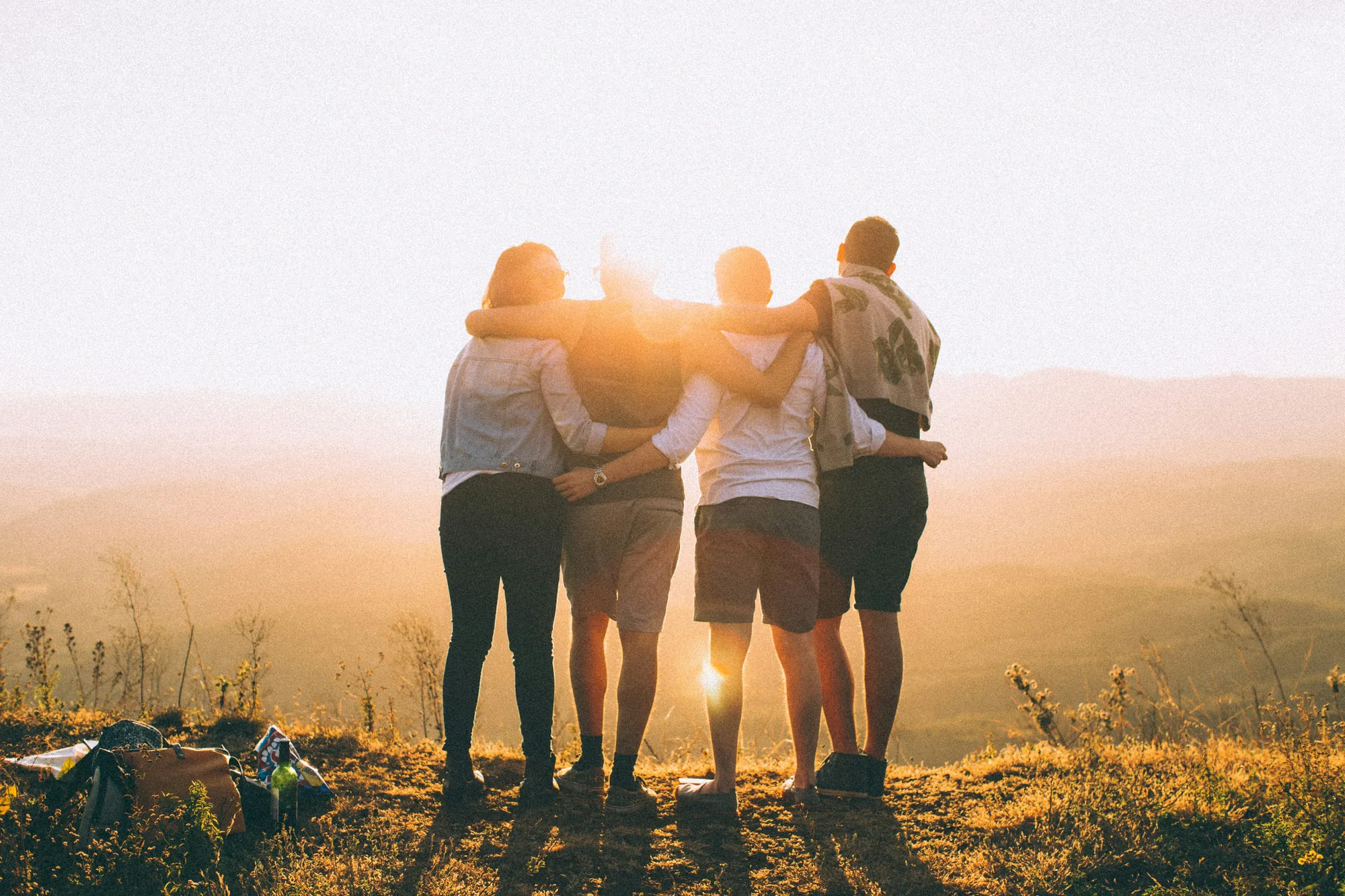 Four friends stand arm in arm on a hillside at golden hour, looking out over the valley together.